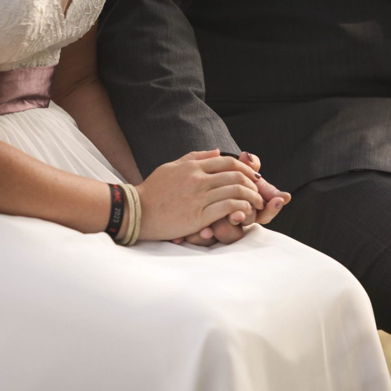Hände eines Brautpaares, die sich liebevoll halten, vor einem Hochzeitskleid.