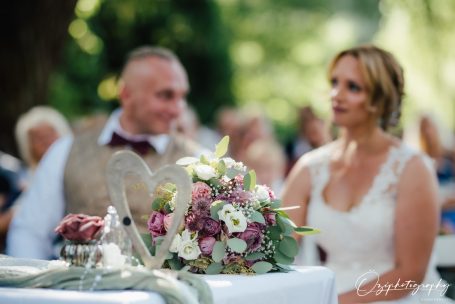 Hochzeitsfeier mit einem Blumenarrangement im Vordergrund und dem Brautpaar im Hintergrund.