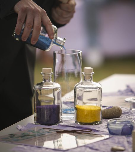 Eine Hand gießt blaue Flüssigkeit aus einer Flasche in ein Glas neben buntem Sand in Gläsern.