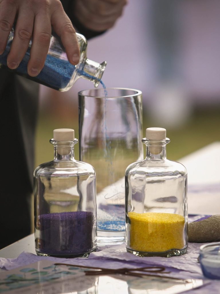 Hände gießen bunten Sand aus Flaschen in ein Glas.