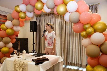 Eine Person steht am Mikrofon vor einem Ballonbogen und einem festlich gedeckten Tisch.