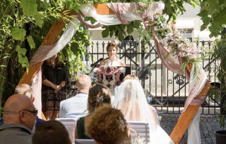 Hochzeitszeremonie im Freien, Braut in weißem Kleid, Gäste im Hintergrund.