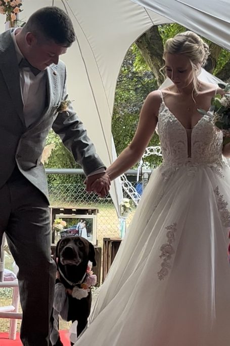 Braut und Bräutigam gehen Hand in Hand zur Feier, ein Hund folgt ihnen.