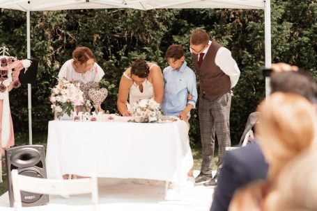 Ein Brautpaar und Gäste unterschreiben Dokumente an einem Hochzeitstisch im Freien.