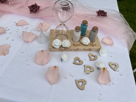 Tischdekoration mit Herzen, Blumen und einem Sanduhr-Set auf einem weißen Tuch.