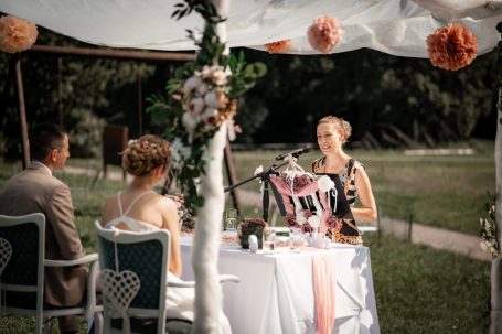 Hochzeitsszene: Brautpaar hört einer Rednerin unter einem Zelt zu.