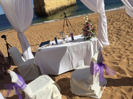 Tisch mit Blumenschmuck und Stühlen am Strand, umgeben von weißen Vorhängen.