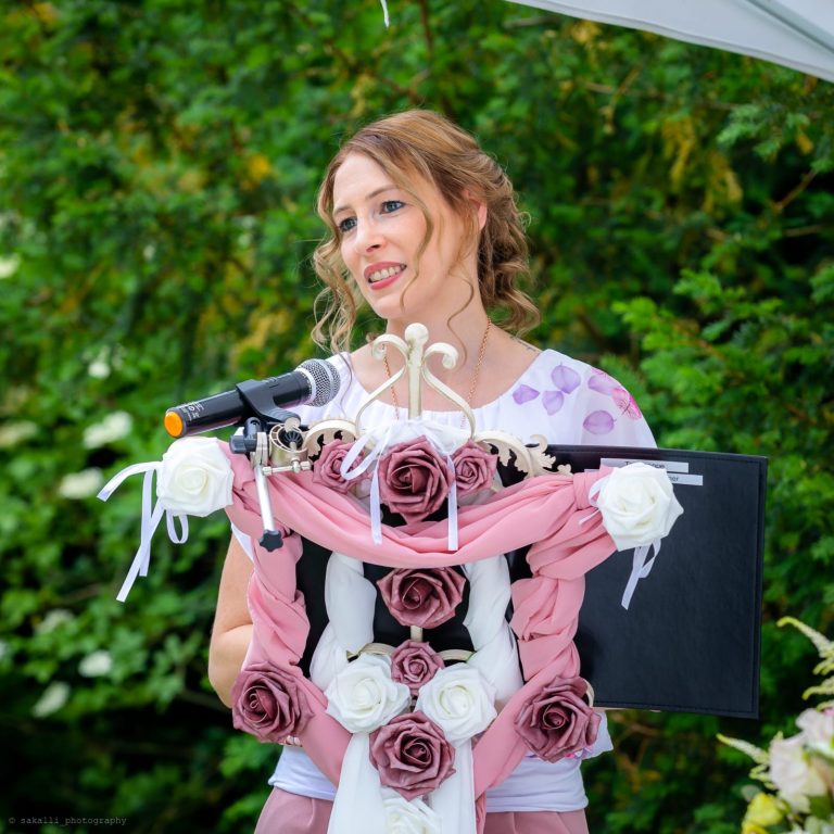 Frau spricht vor einem Mikrofon, umgeben von Rosen und Grünpflanzen.
