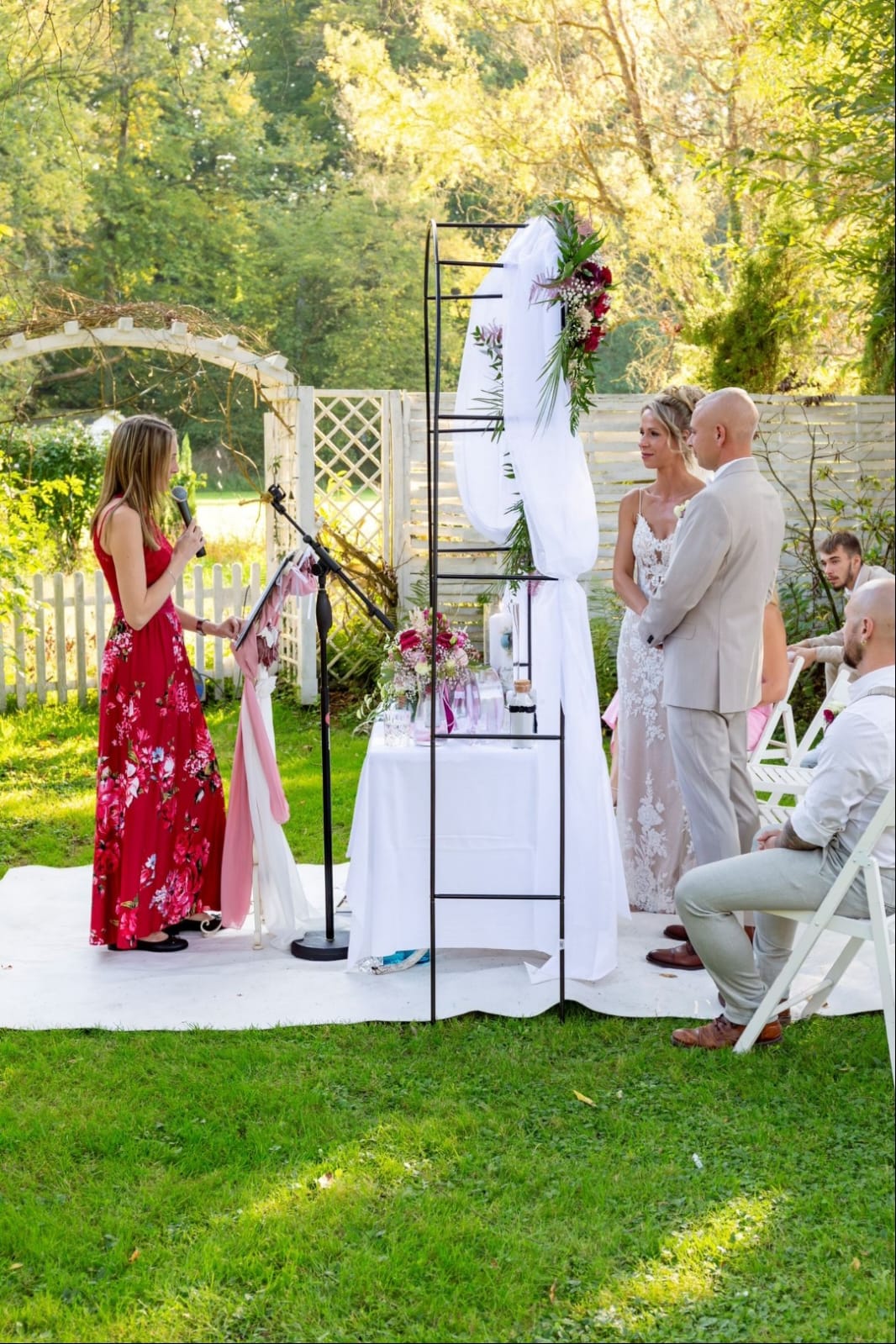 Eine Hochzeitszeremonie im Garten mit Brautpaar und Rednerin vor dekoriertem Hintergrund.