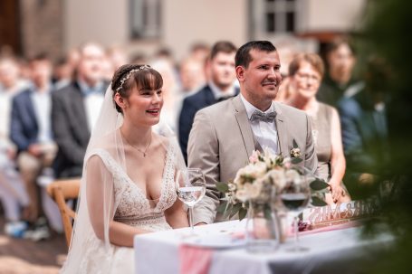 Ein lächelndes Brautpaar sitzt am Hochzeitstisch, umgeben von Gästen.